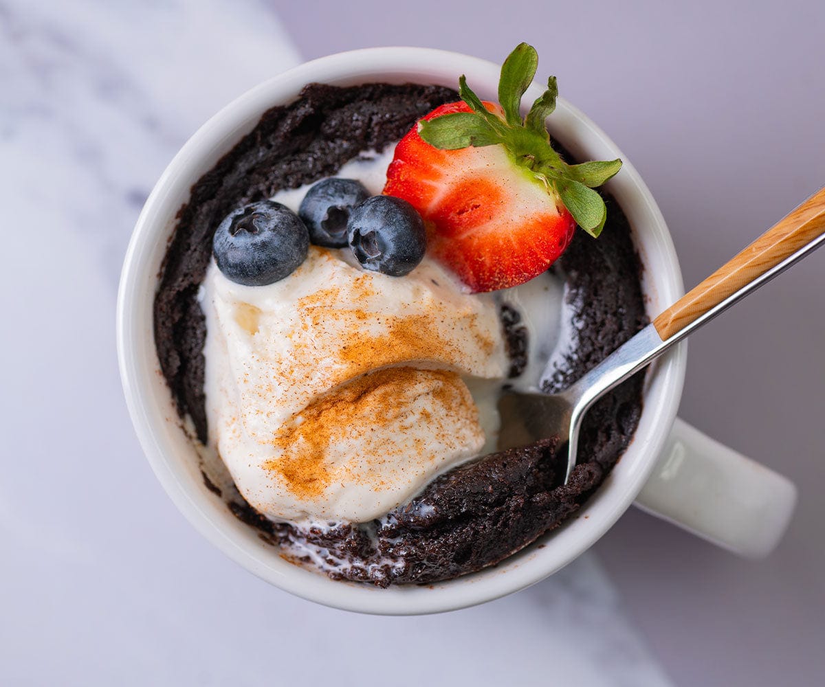 Overhead shot of a finished mug brownie with a spoon, ice cream, and fresh blueberries and half a strawberry