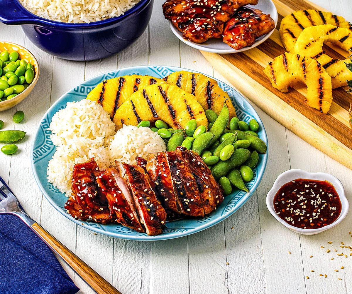 Finished dish of Huli Huli Chicken with race, grilled pineapple, and edemame on a table with more chicken, sauce, pineapple and rice