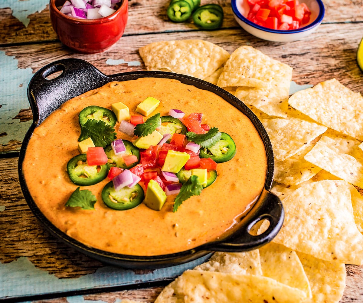 Finished skillet of Tex-Mex Queso with chips garnished with onion, tomatoe, avocado, and jalapeno slices